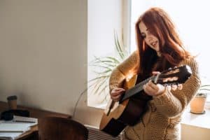 Indoor cozy Hobbies for winter, autumn cold season. Redhead woman playing acoustic guitar and listening to music, sitting near window home